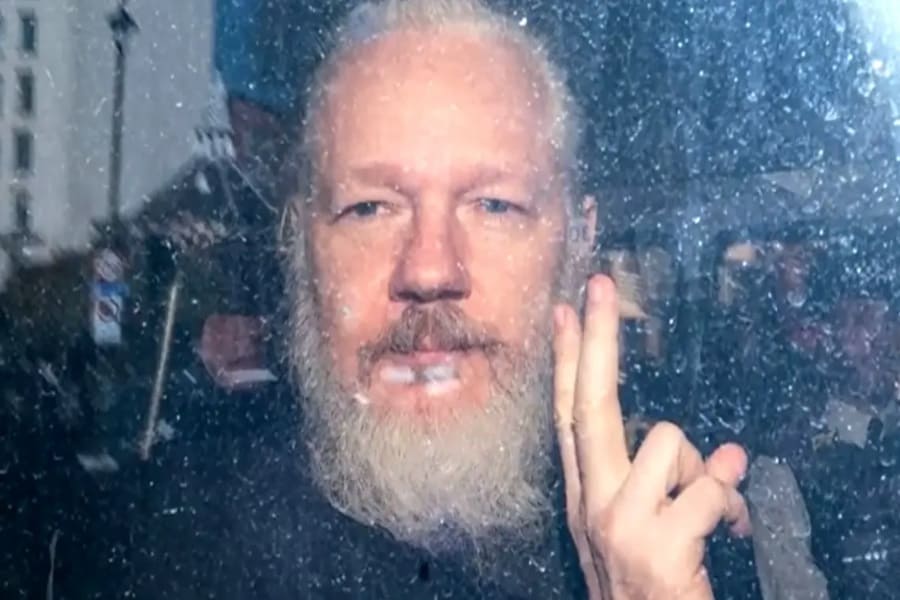 Julian Assange saluda a las cámaras con barba crecida, a través de un cristal.