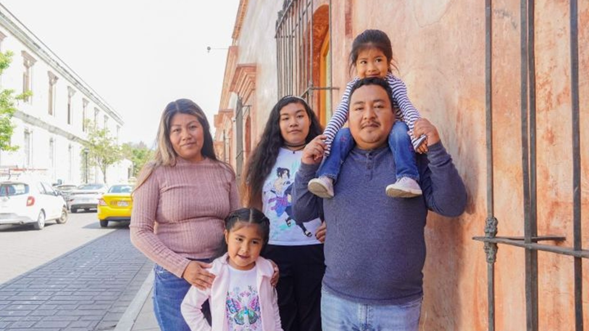 Un hombre carga a una niña a hombros, junto a otra niña de pie, una mujer joven y una más adulta, en una calle.