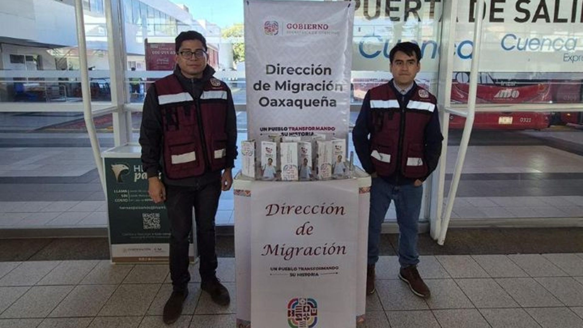 Dos personas con chaleco guinda paradas junto a un stand informativo.