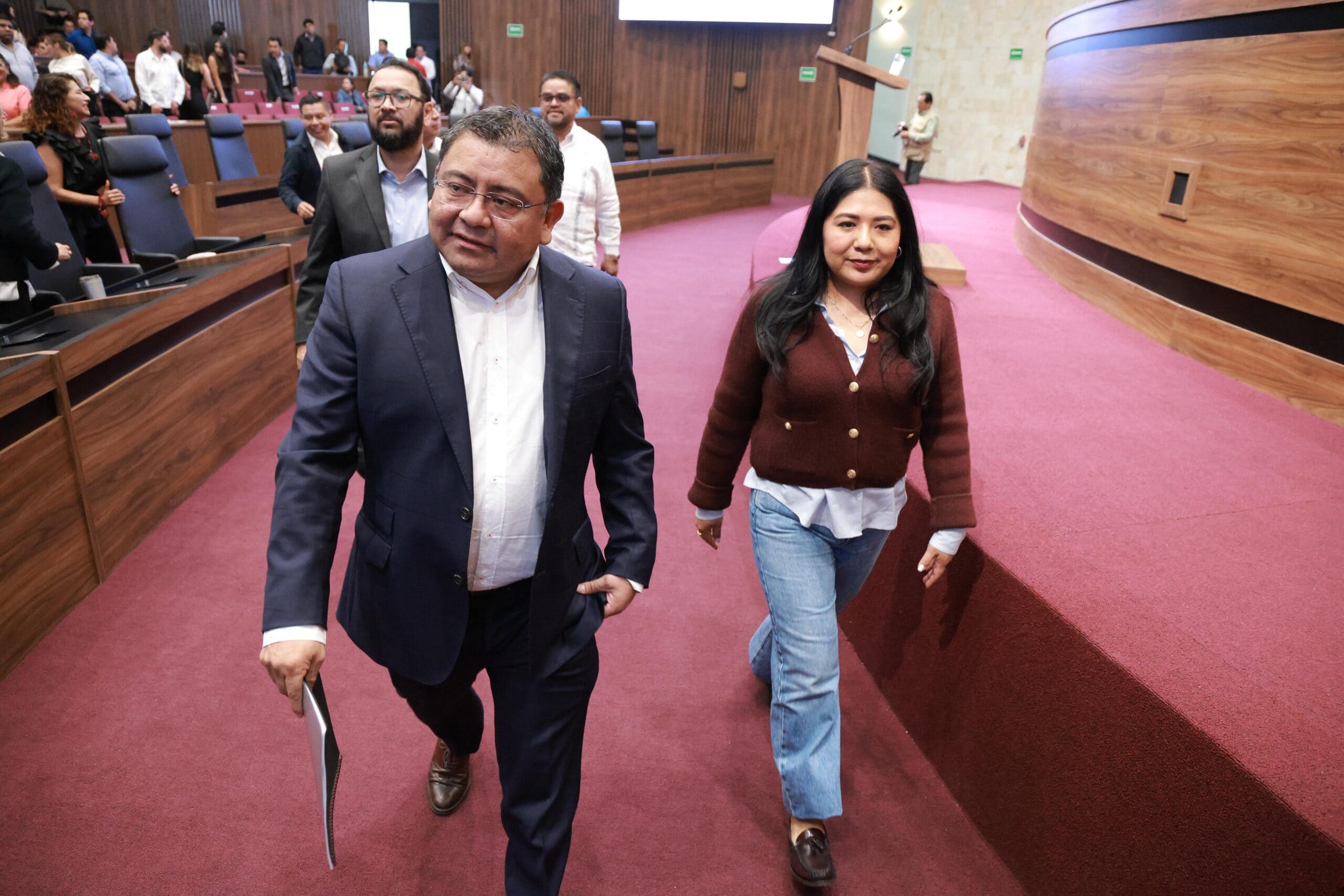Un hombre con traje negro y una mujer con blusa roja caminan entre un numero grupo de personas.