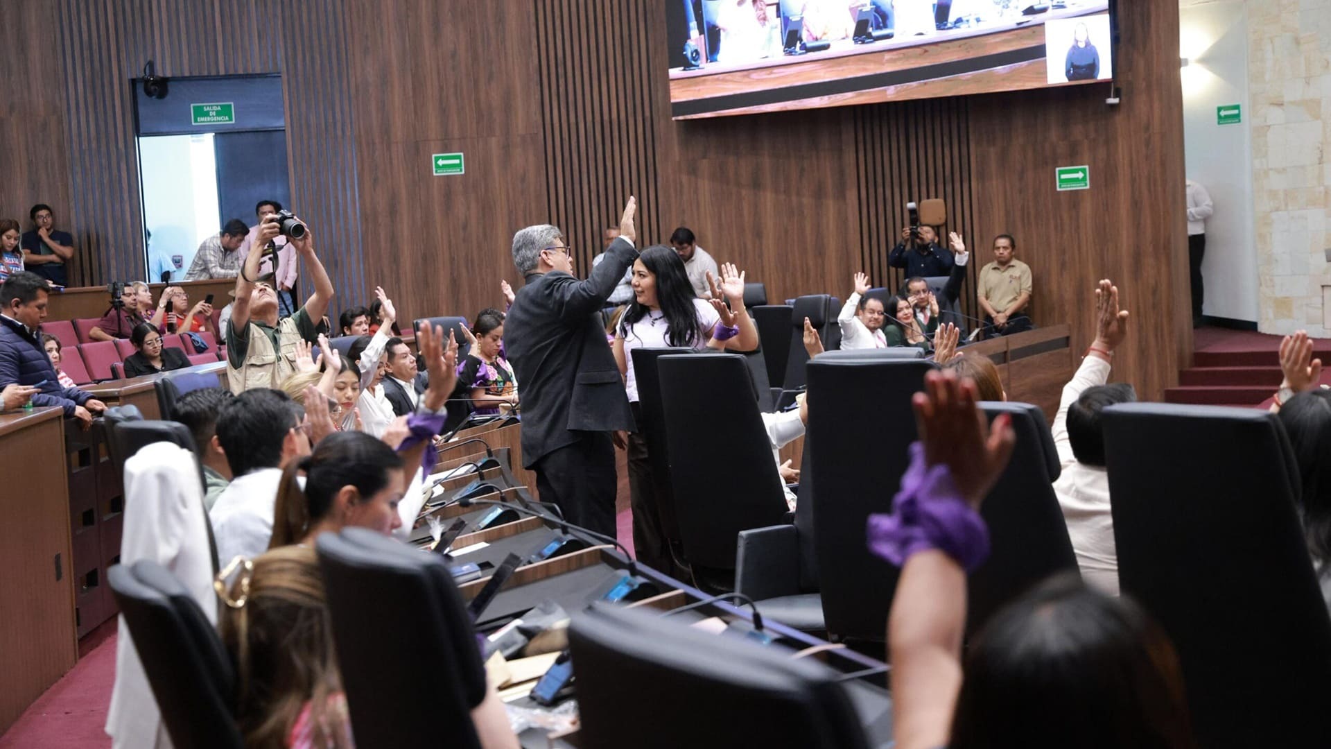Un grupo de personas reunidas en un gran salón levantan la mano al mismo tiempo.