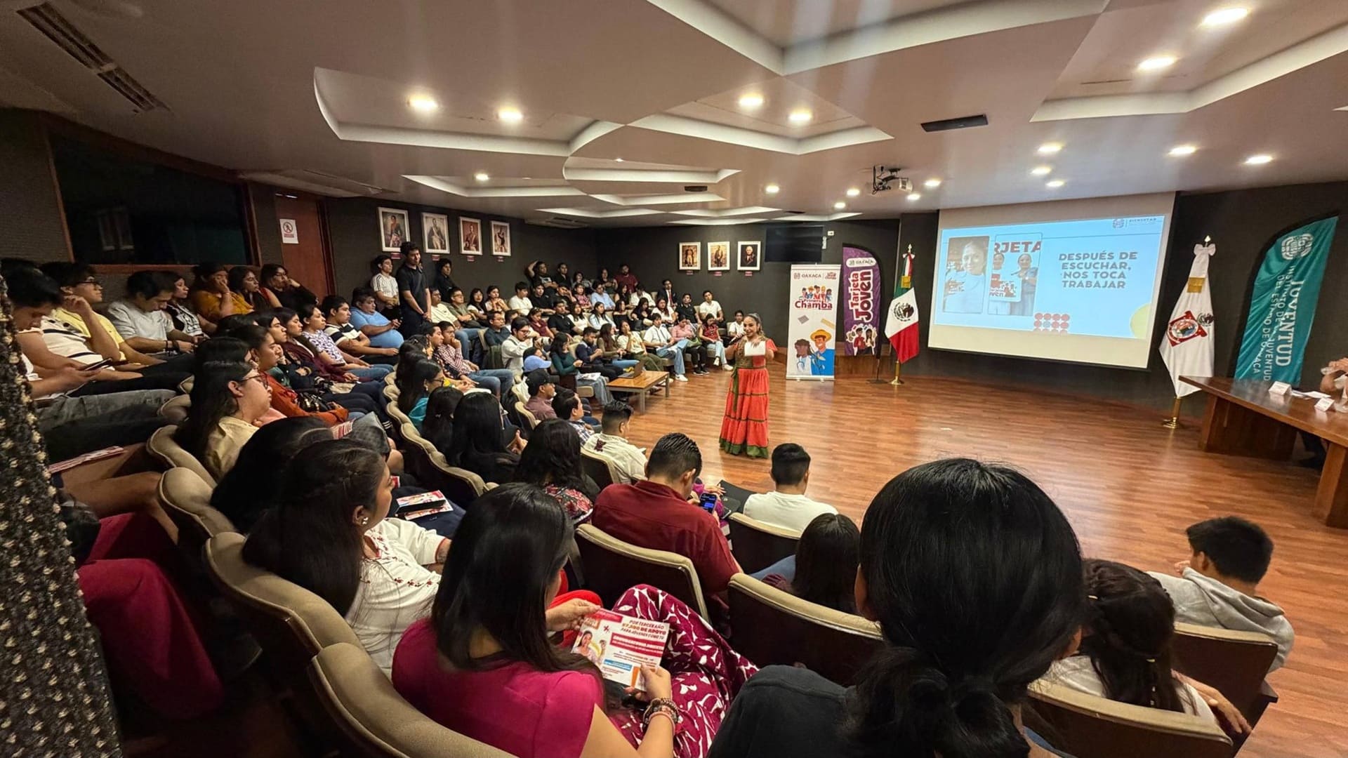 Mujer vestida con traje típico habla en un auditorio lleno de jóvenes.