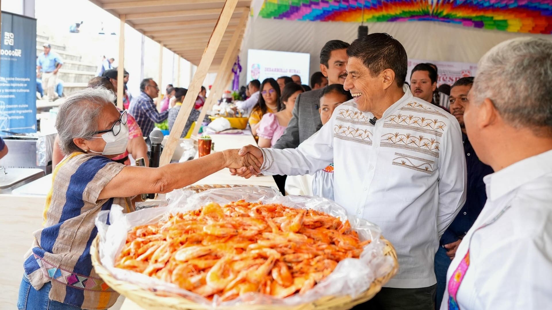 Un hombre con camisa blanca saludando a una afromexicana junto a una canasta con camarones secos.
