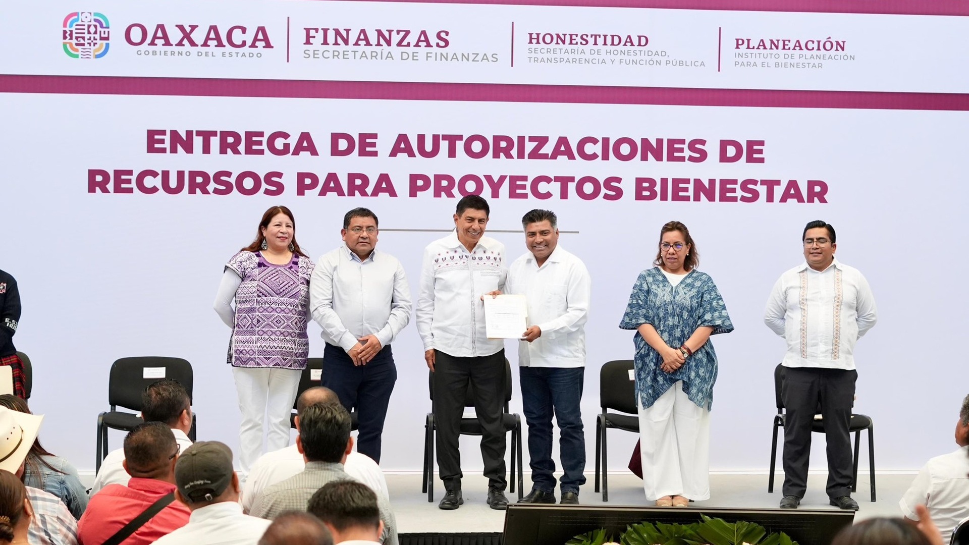Cuatro hombres y dos mujeres en el templete, uno de ellos mostrando una hoja y siendo retratados.