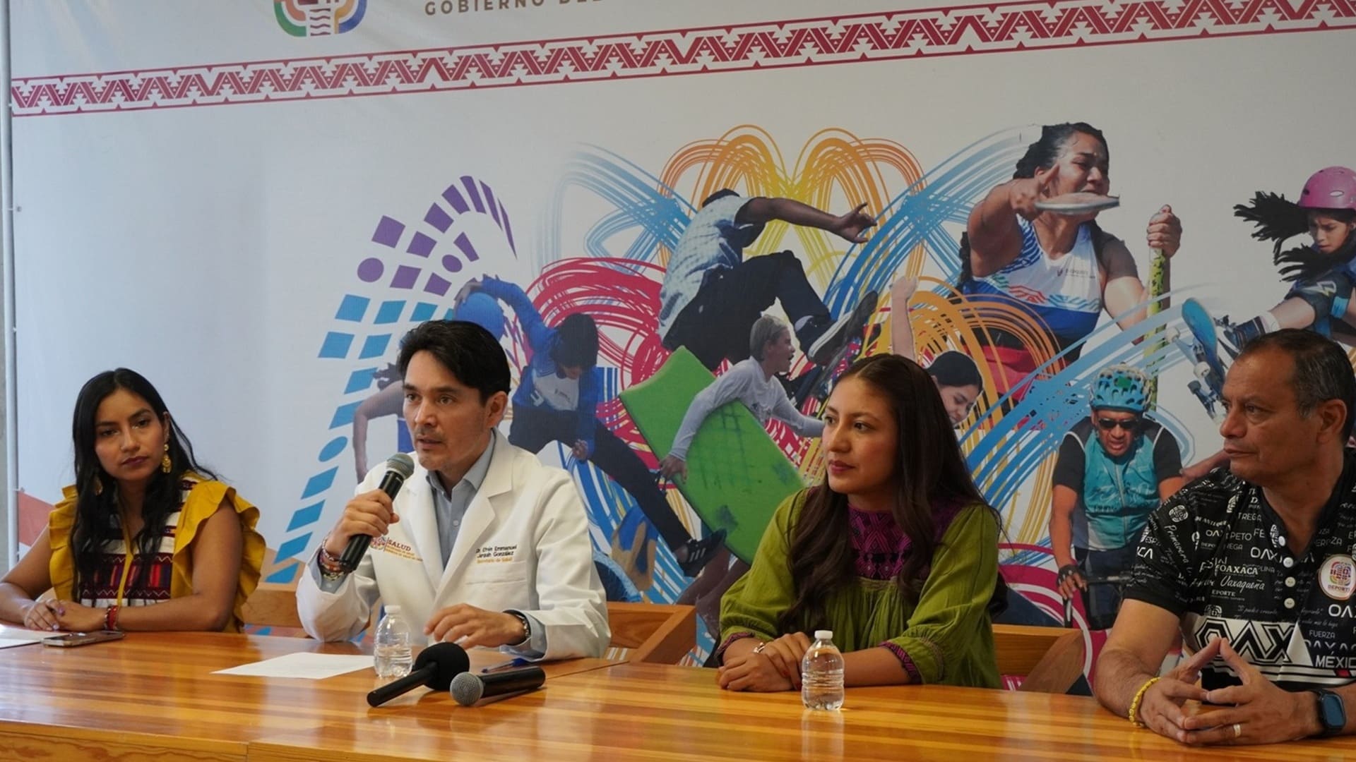 Un hombre con bata blanca habla con un micrófono, junto a tres mujeres también sentadas.