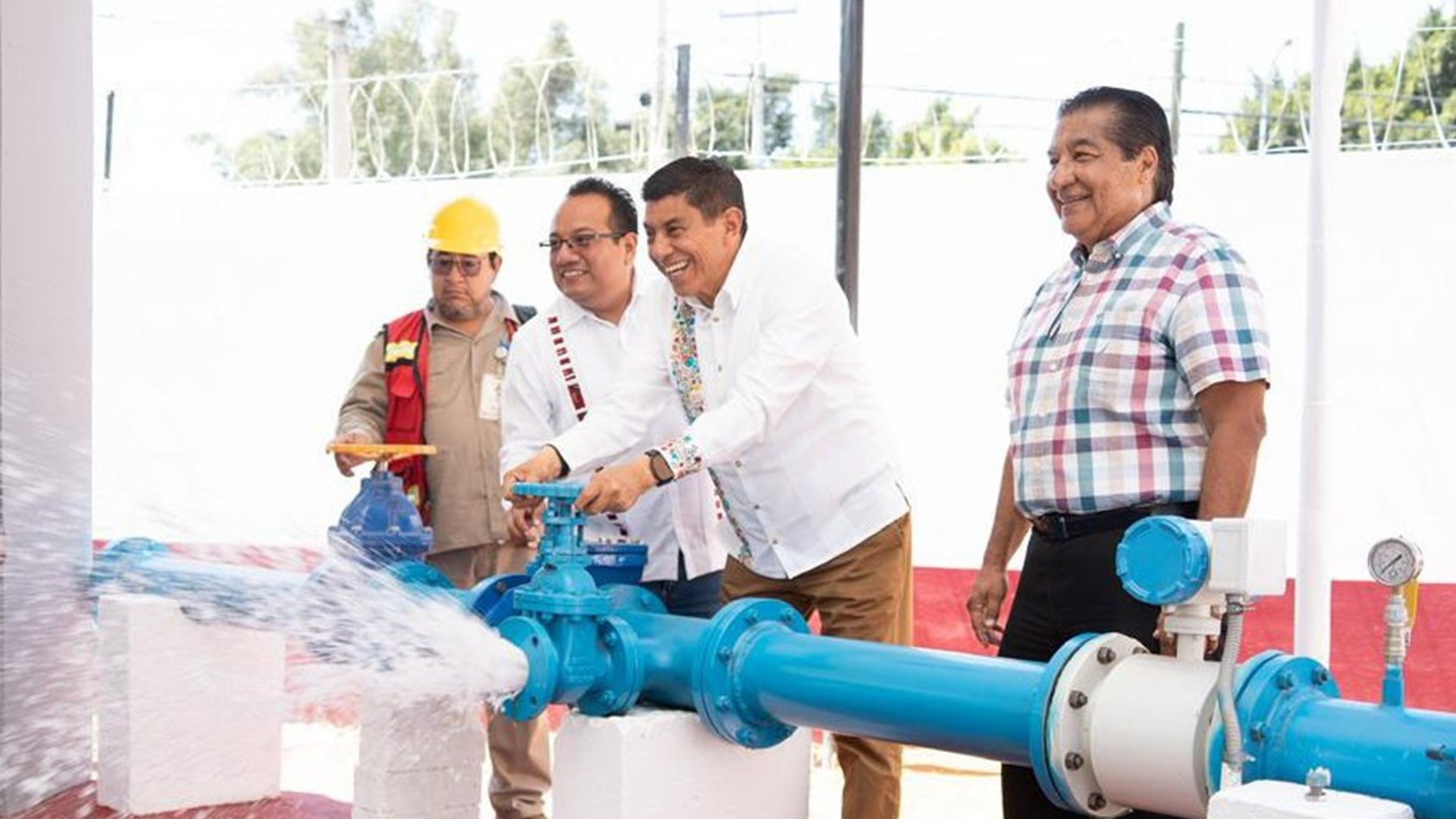 Cuatro hombres de pie, uno con casco de obrero y uno manipula una bomba de agua que avienta un chorro.