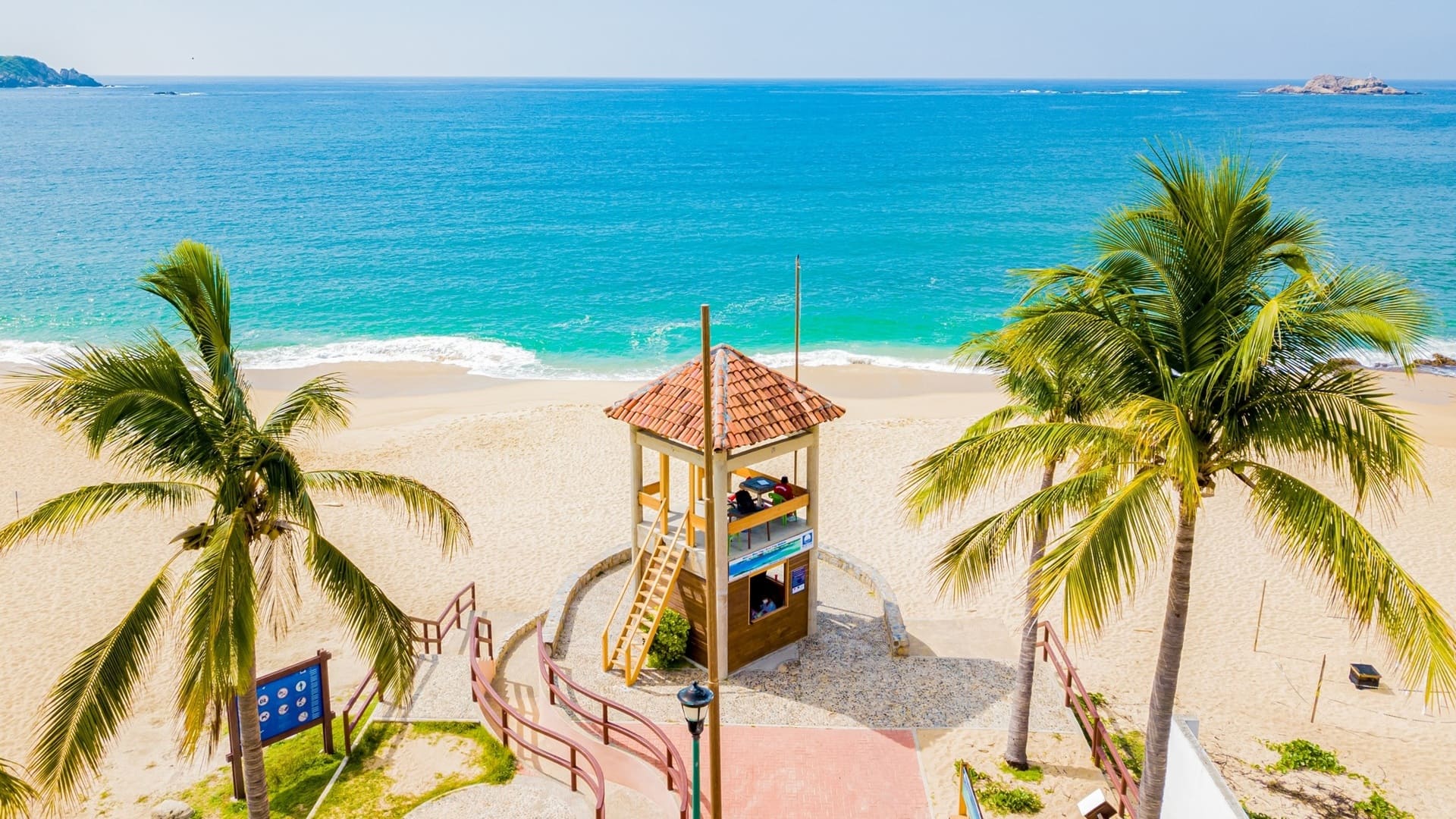 Imagen de una playa con agua azul intenso, dos palmeras y una torre de vigilancia.