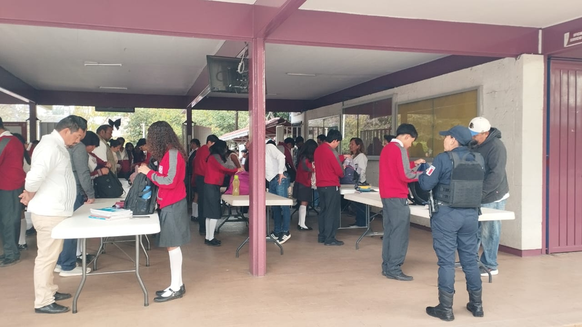 Personas realizan la revisión de mochilas de alumnos a la entrada de una escuela, con la vigilancia de policías.