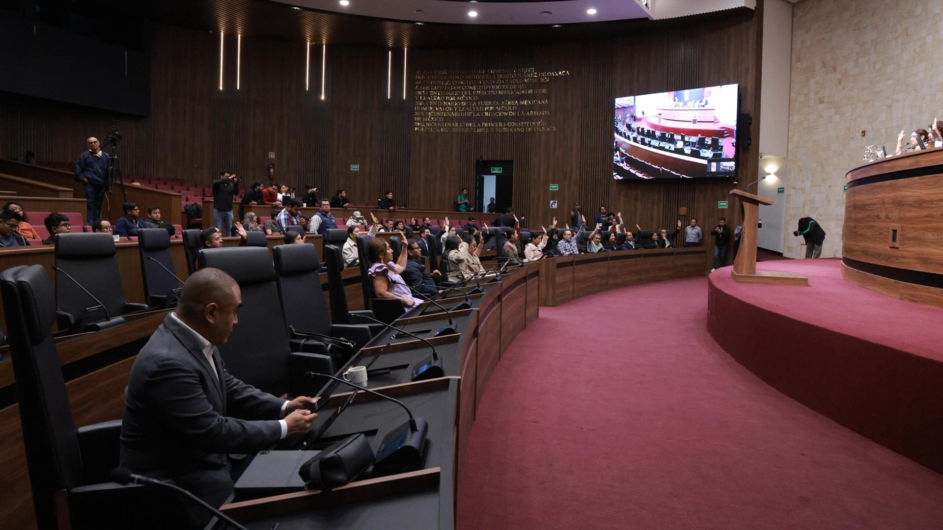 Vista general del Congreso de Oaxaca con algunos hombres y mujeres sentados en sus asientos en una sala con alfombra roja.