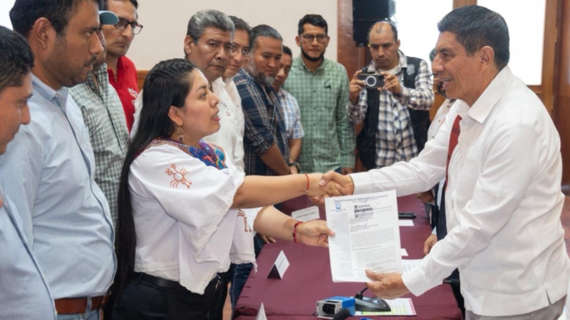 Salomón Jara saluda a una mujer, rodeado de muchas otras y exhibe un documento sobre una mesa.