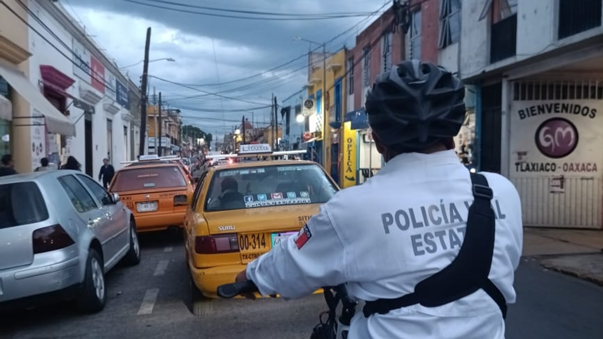 Policía vial de Oaxaca con casco y en una bicicleta recorre una calle atrás de un taxi amarillo y otros autos.