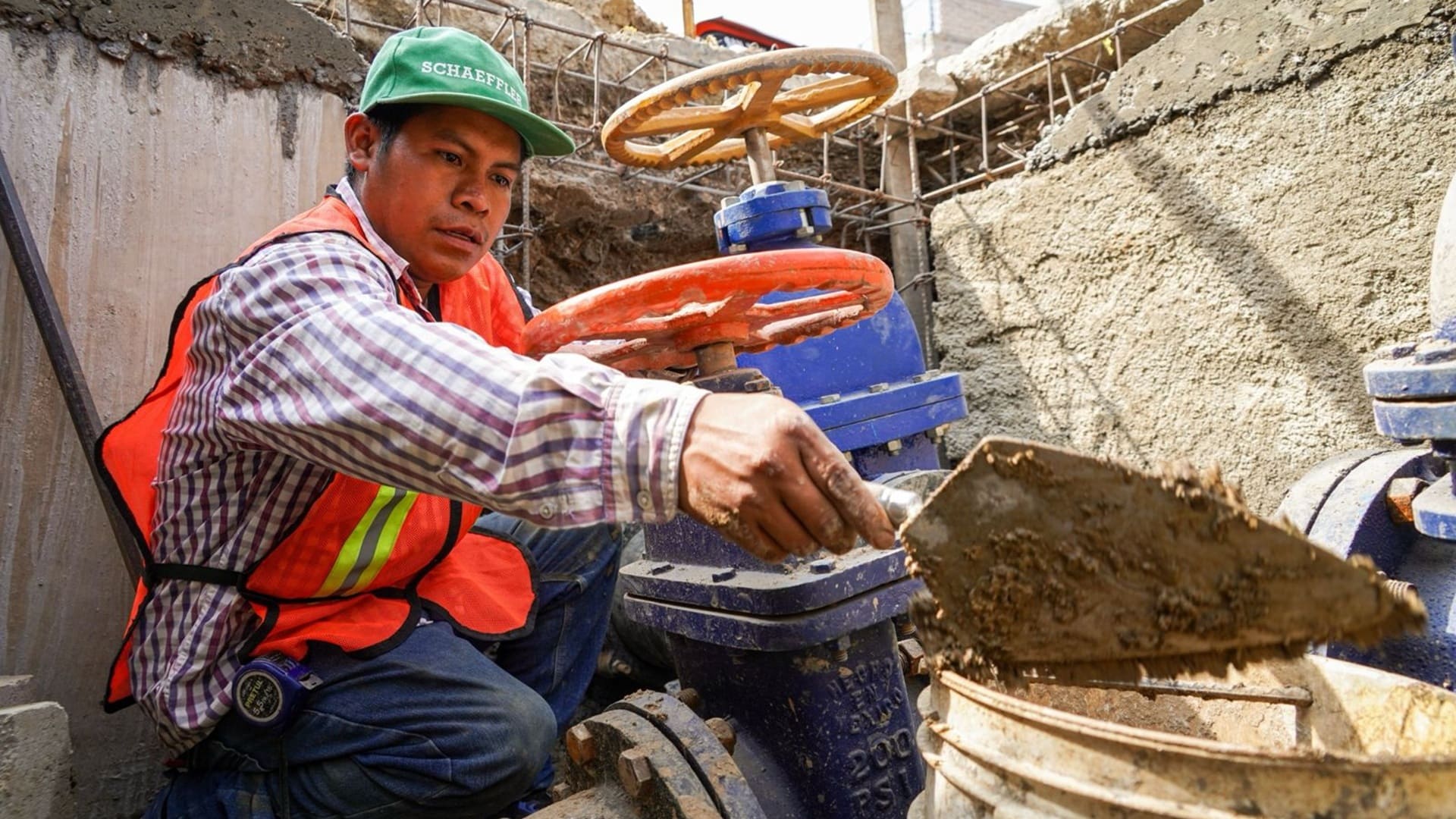 Trabajador con chaleco naranja y gorra verde manipulando una cuchara con la que lleva tierra a una cubeta.