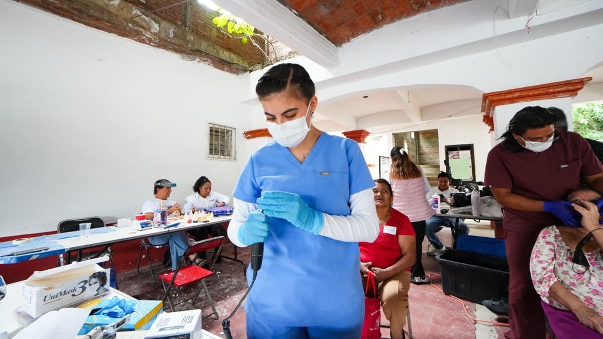 Trabajadora de la salud dental con uniforme azul y tapabocas colocándose guantes en una sala donde hay personas sentadas