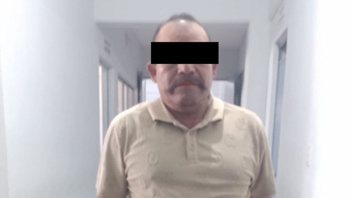 Persona vestido de beige con los ojos cubiertos por ley posa frente a la cámara con las manos esposadas