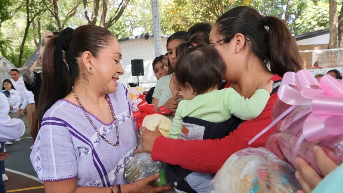 Irma Bolaños, vestida con huipil morado, entrega una bolsa con productos a una mujer que lleva un bebé en brazos
