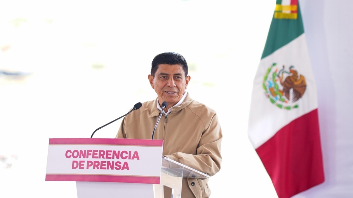 Salomón Jara con chamarra café y junto a la bandera de México ofreciendo una conferencia de prensa