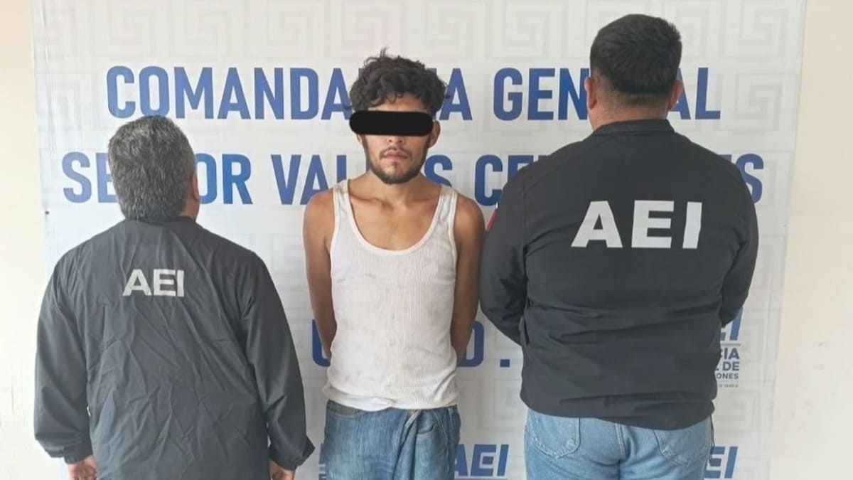 Hombre joven con camiseta blanca, esposado y con una franja negra sobre los ojos por mandato de ley; custodiado por dos elementos de seguridad pública