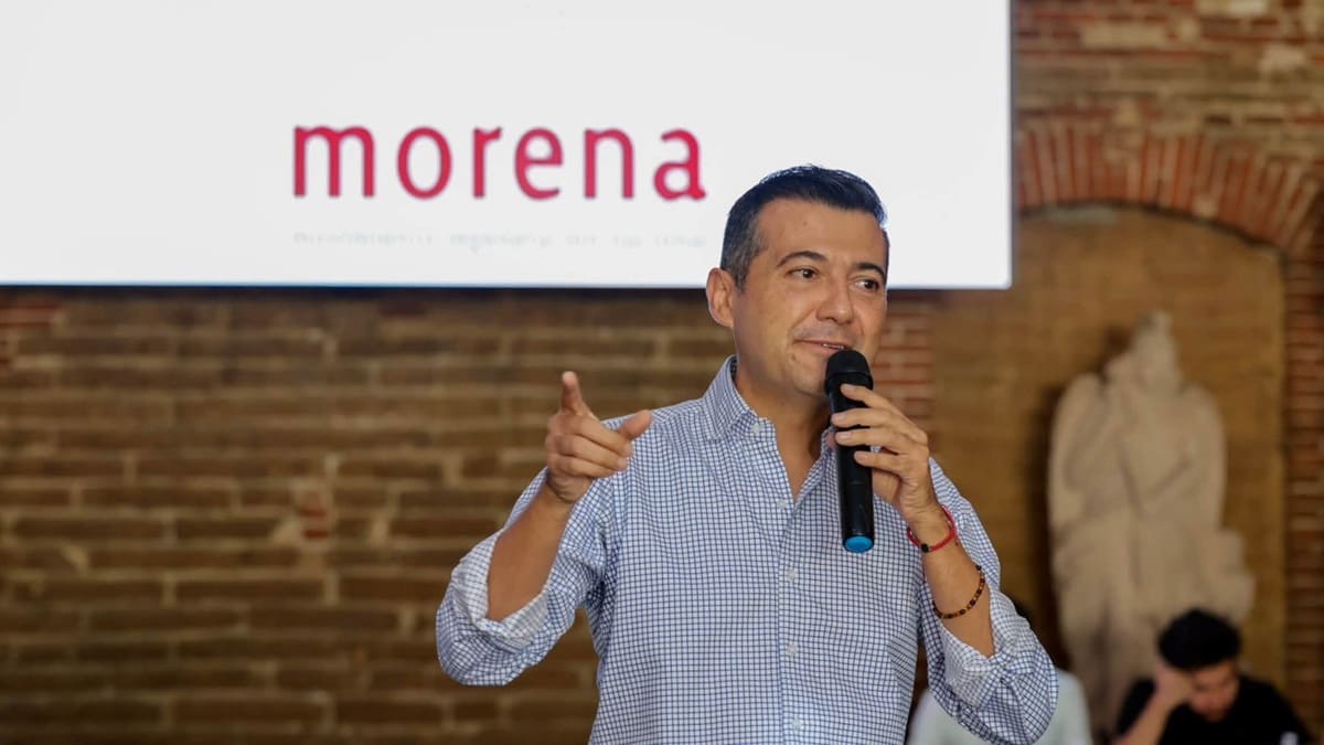 Luis Alfonso Silva Romo con micrófono en la mano y junto a un letrero de MORENA