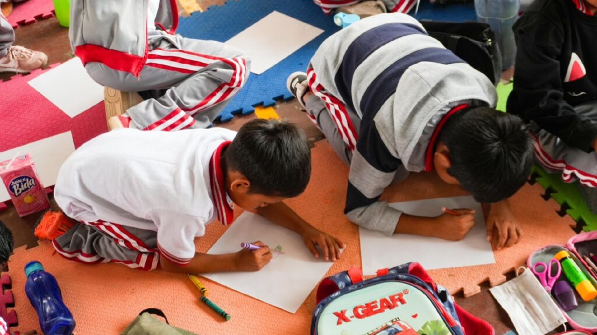 Dos niños sentados en el piso escriben sobre hojas en blanco, rodeados de artículos escolares.