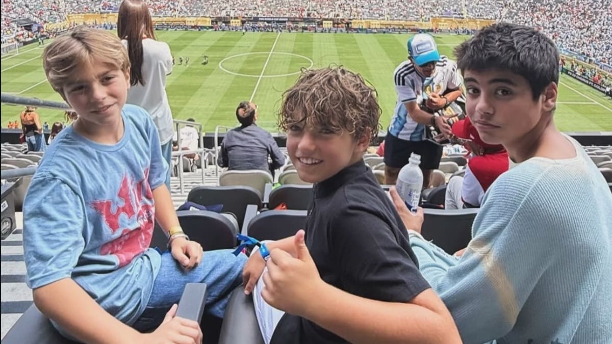 Tres jóvenes entre ellos dos hijos de Shakira, saludan a la cámara sentados en la tribuna del estadio de Nueva Jersey