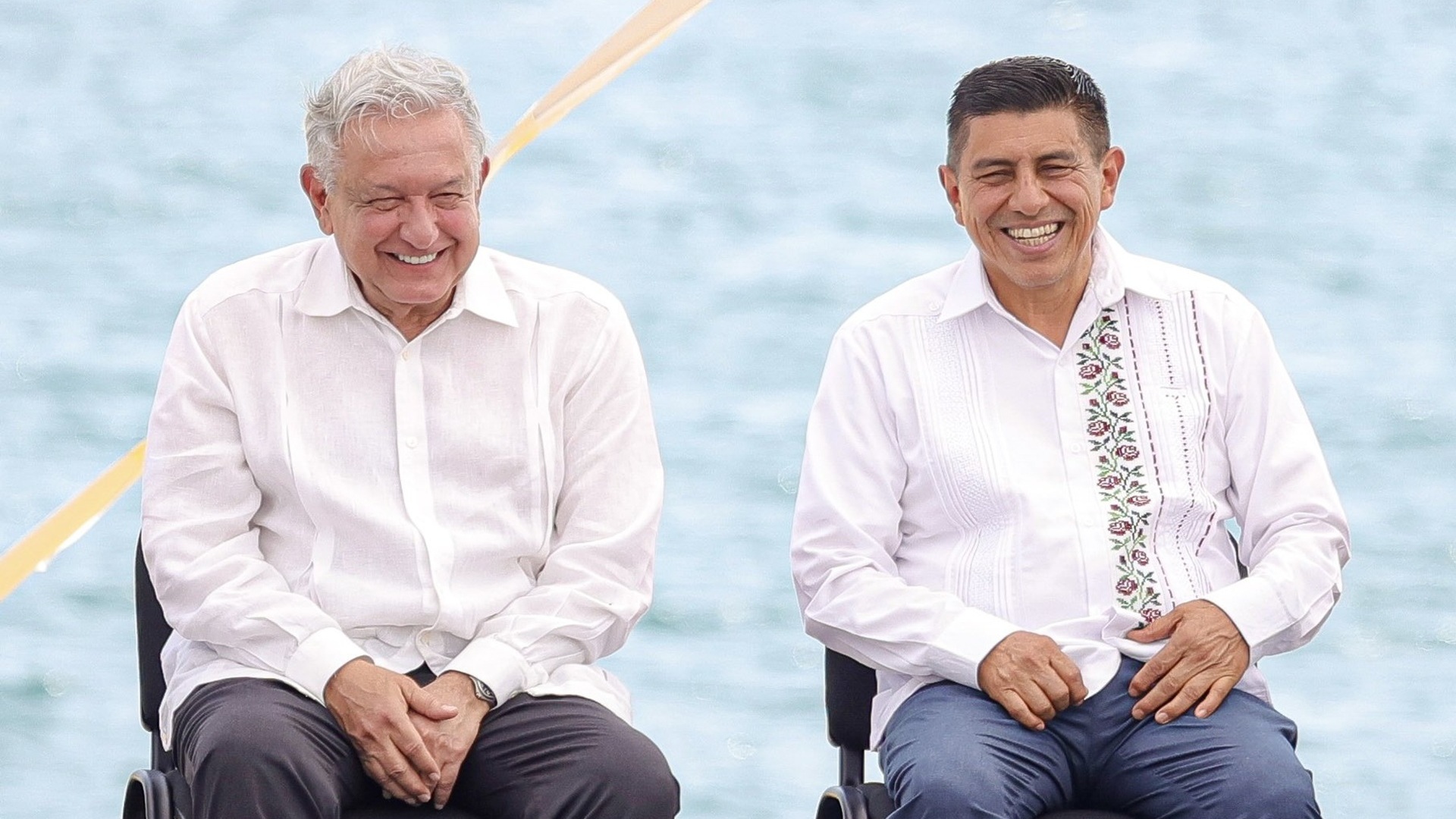 AMLO y Jara Cruz sentados frente  mar y sonriendo a la cámara.