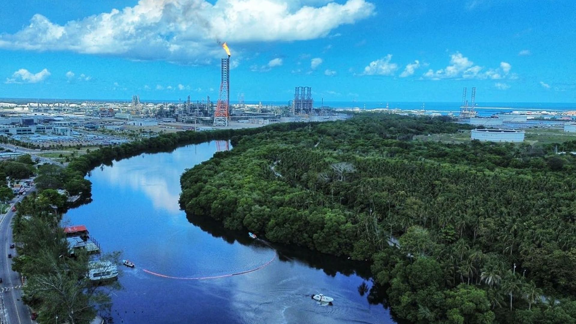 Imagen aérea de un río, con abundante vegetación a los costados y una refinería al fondo.
