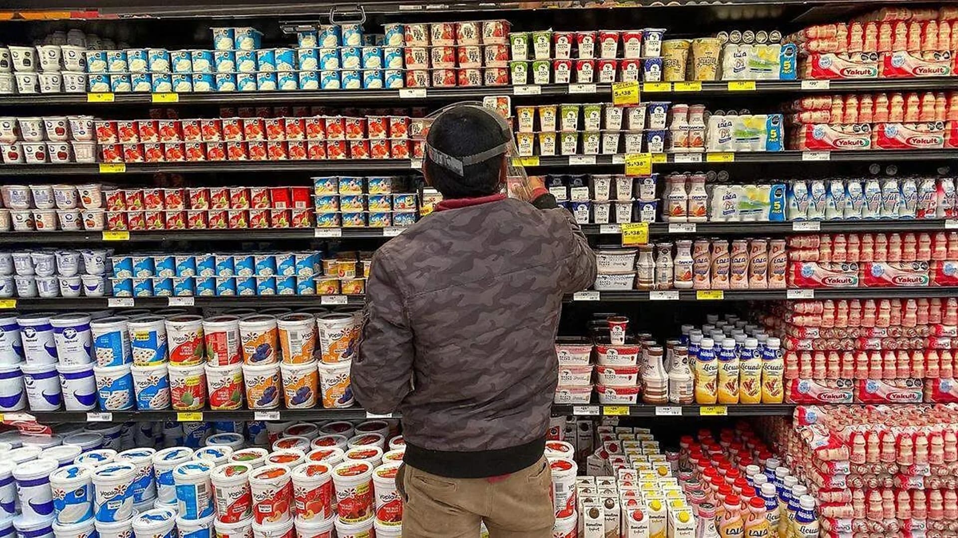 Un hombre con chamarra y pantalón café observa los precios de los productos en el refrigerador de un centro comercial.