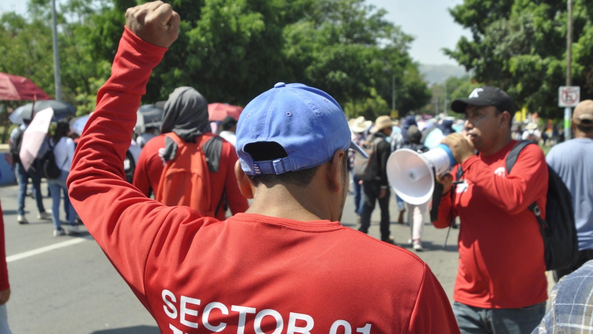 Un hombre con sudadera roja y gorra azul levanta la mano en señal de protesta junto a otro con un altavoz.