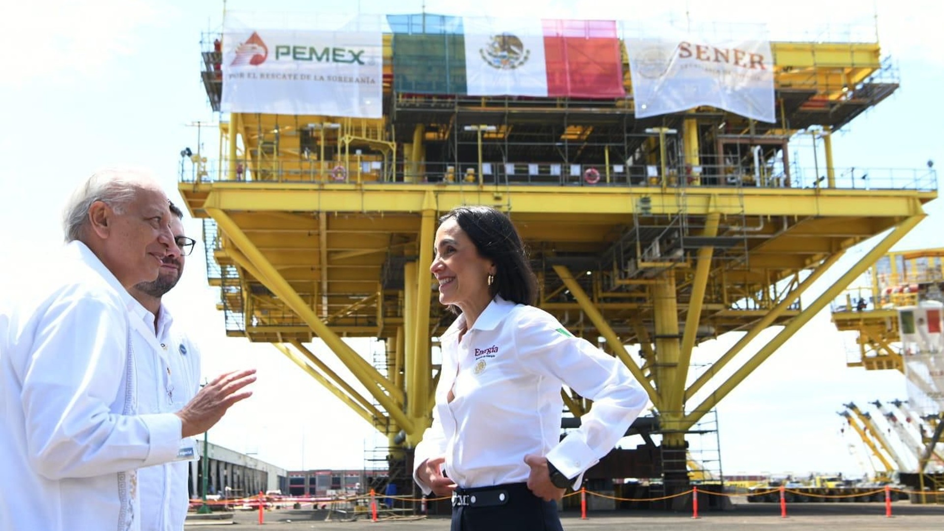 Dos hombres y una mujer, funcionarios de Energia y Pemex, con camisas blancas platicando frente a instalaciones de gran tamaño.