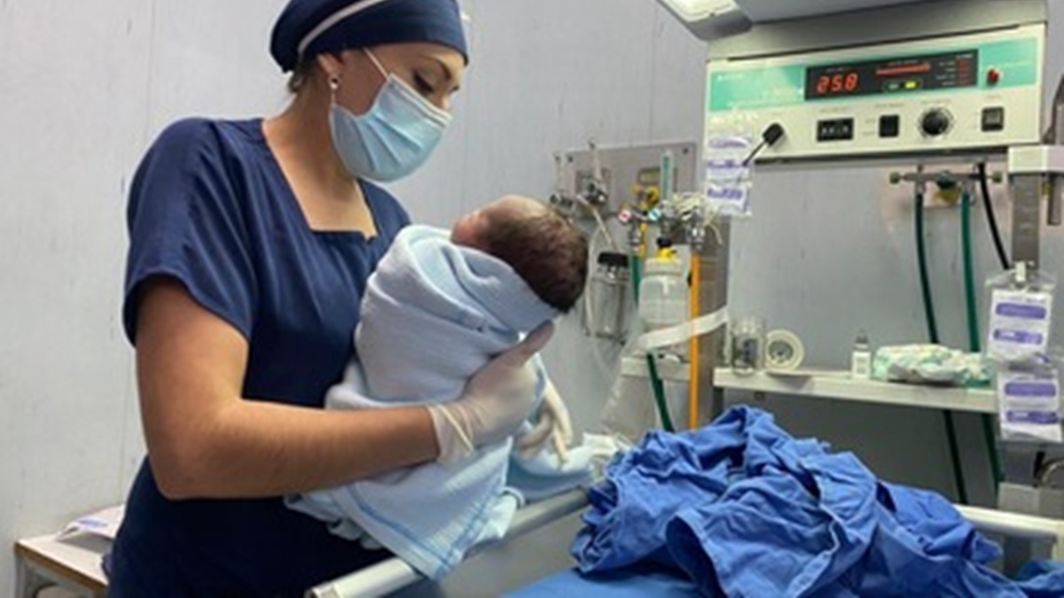 Enfermera con uniforme azul y tapabocas carga a un bebé recién nacido 