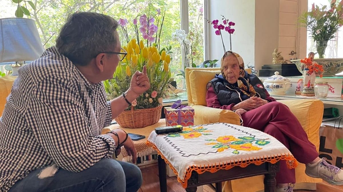 Elena Poniatowska sentada en una biblioteca y con pañoleta en la cabeza, en plática con Domingo García Mijangos, vestido con camisa azul de cuadros