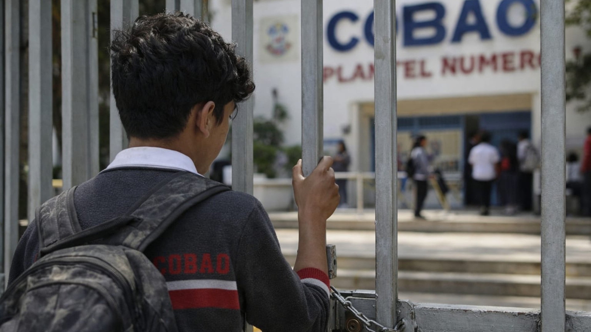 Un niño con mochila detrás de una reja y mirando una entrada con el letrero "Cobao".