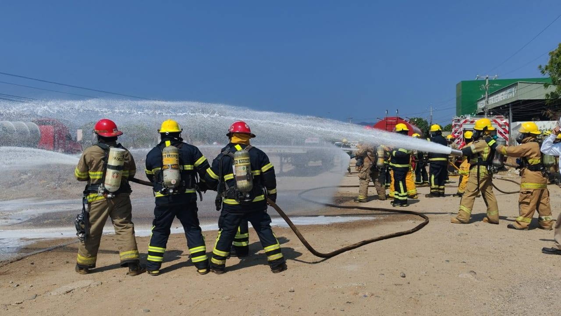 Dos grupos de bomberos con uniforme y casco sostienen manguera mientras lanzan chorros de agua.