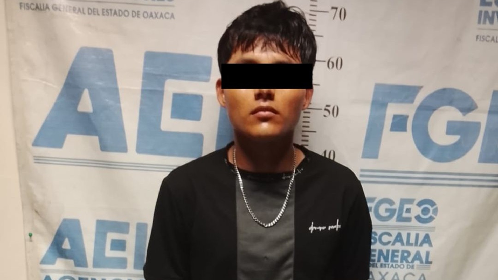 Hombre joven vestido de negro y con una cadena posa con el rostro protegido y frente a una lona de la FGEO.