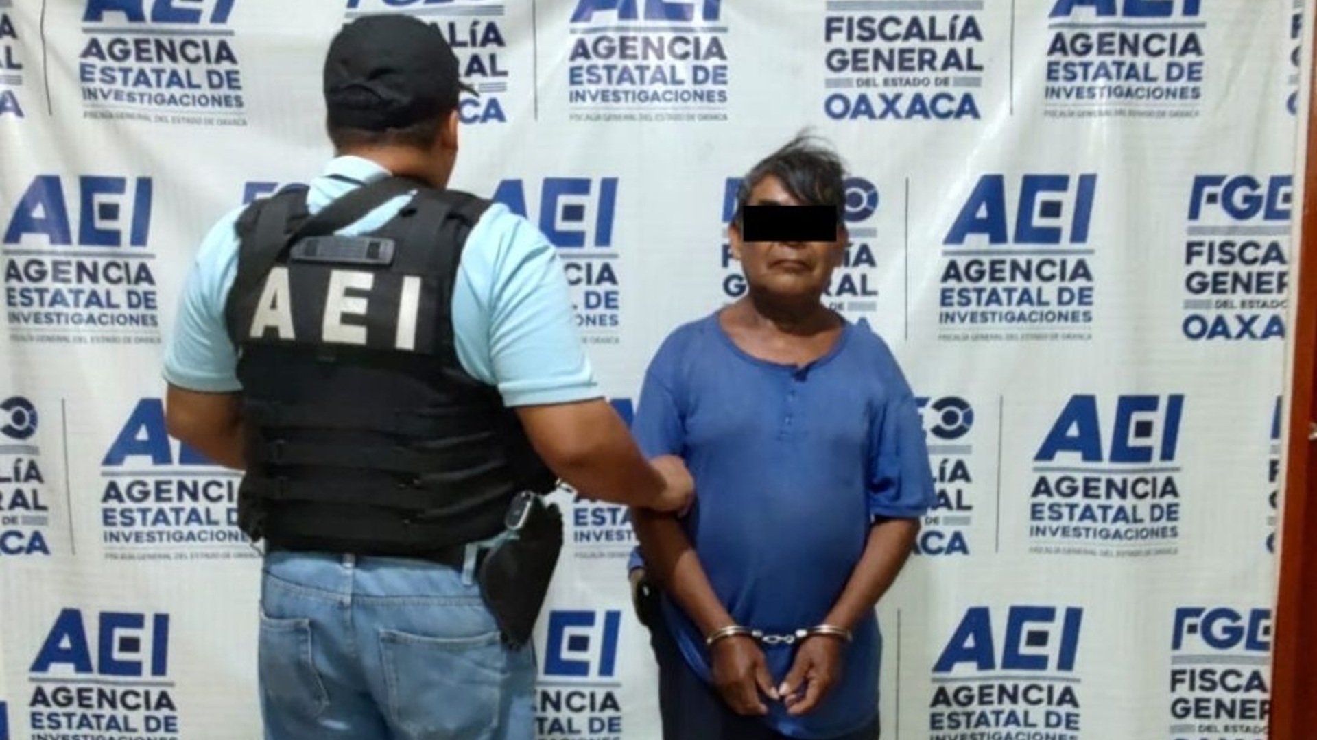 Un hombre con playera azul y esposas, con el rostro protegido, junto a un agente de la AEI.