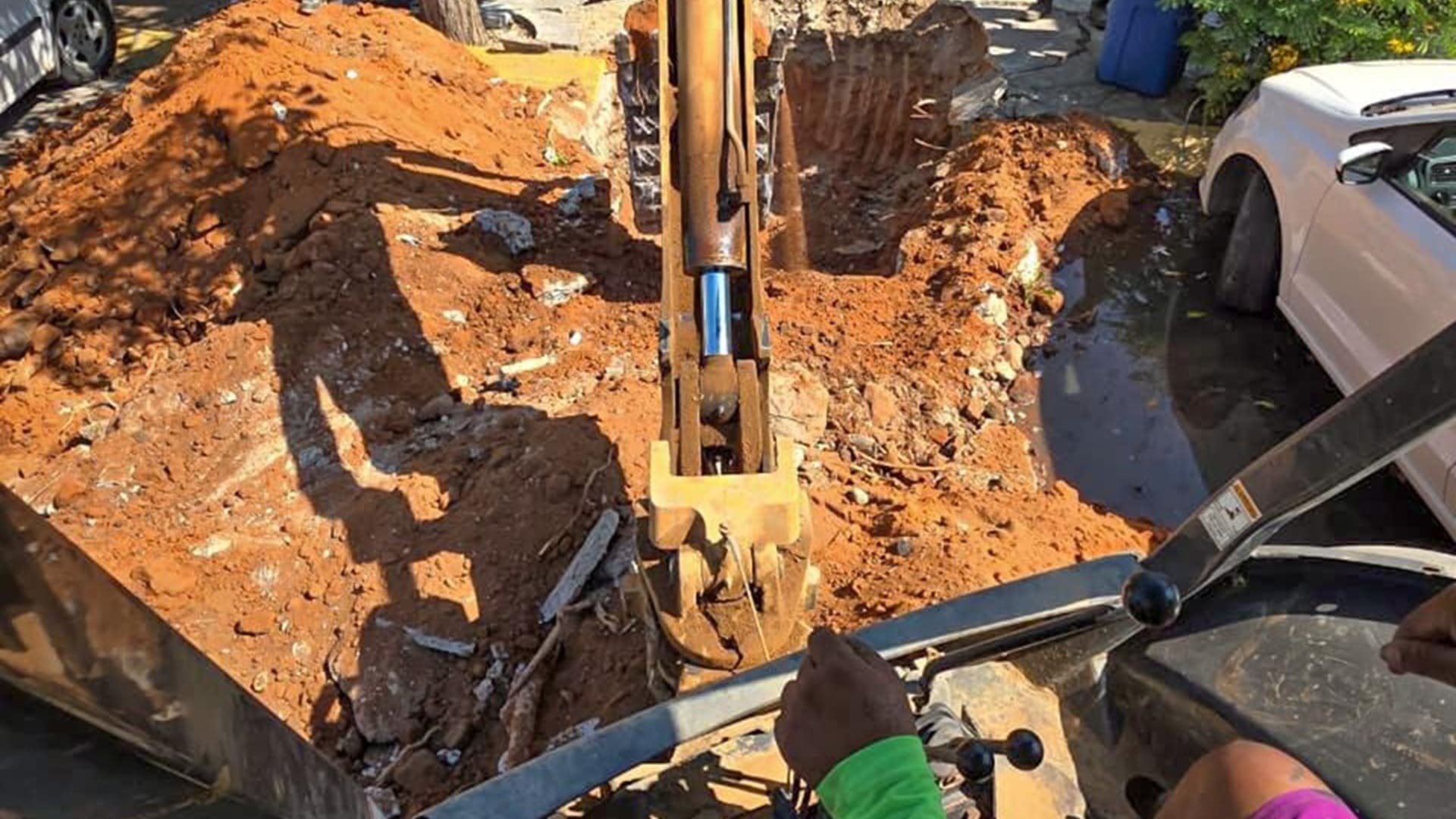 Una persona manipula una máquina excavadora mientras realiza un hoyo en la tierra, junto a un auto blanco.