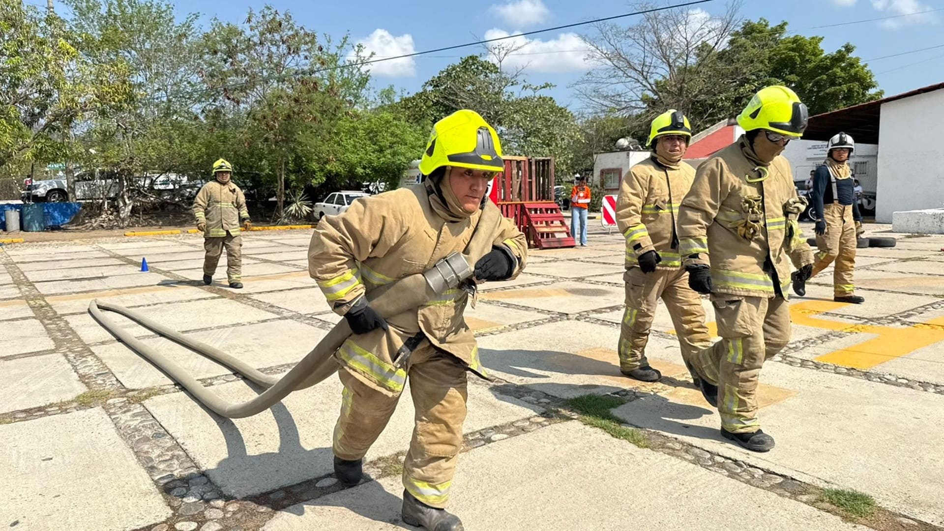 varios bomberos con uniforme y casco amarillo arrastran mangueras en un patio exterior.