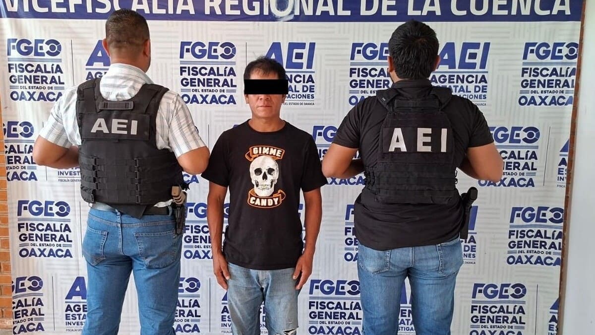Hombre con camisa negra, rostro limpio, jeans y ojos cubiertos por ley, custodiado por dos elementos de la AEI