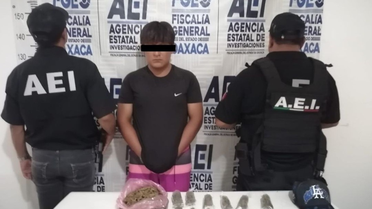 Persona joven con playera negra y pantalón rosa y ojos cubiertos por ley, custodiado por dos agentes y frente a una mesa con droga