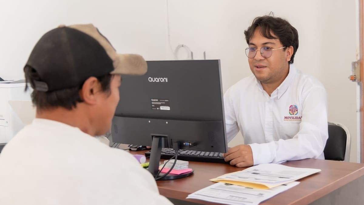 Hombre vestido con camisa blanca frente a una computadora conversar con otro que usa gorras y sentado, quien escucha indicaciones